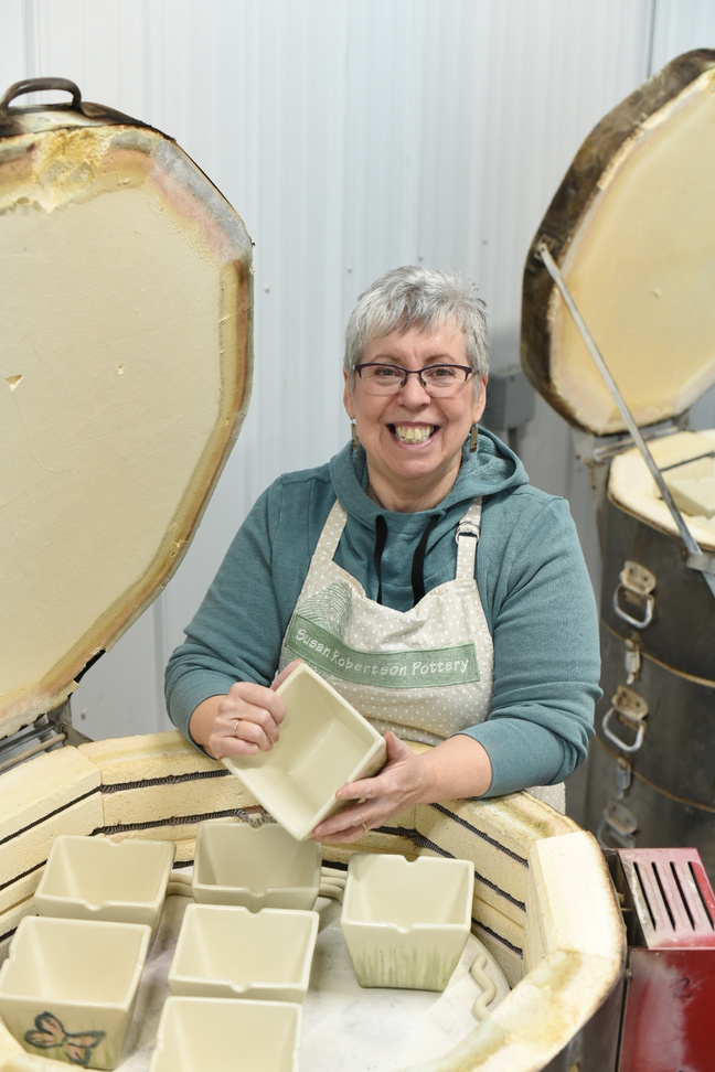 A photo of Canadian ceramic artist Susan Robertson standing next to an open kiln. She is holding some pottery in her hands and smiling.